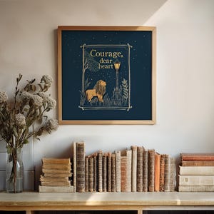 May include: A framed art print with a navy blue background features a lion illustration and the words "Courage, dear heart" in gold lettering. The artwork is in a wooden frame and is displayed above a collection of vintage books.