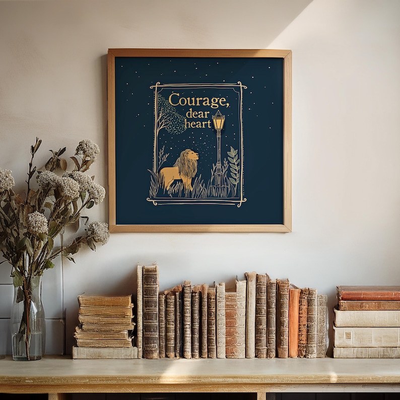 May include: A framed art print with a navy blue background features a lion illustration and the words "Courage, dear heart" in gold lettering. The artwork is in a wooden frame and is displayed above a collection of vintage books.