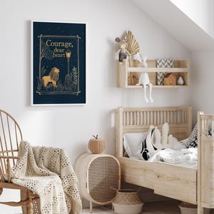 May include: A framed art print with a navy blue background features a lion illustration and the words "Courage, dear heart" in gold. The artwork is displayed in a child's bedroom with a wooden bed and decorative shelf.