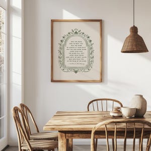 May include: A framed print with a wooden frame, featuring a green floral border and text. The text reads "May the Road Rise to Meet You." The print is displayed in a dining room setting with a wooden table and chairs.