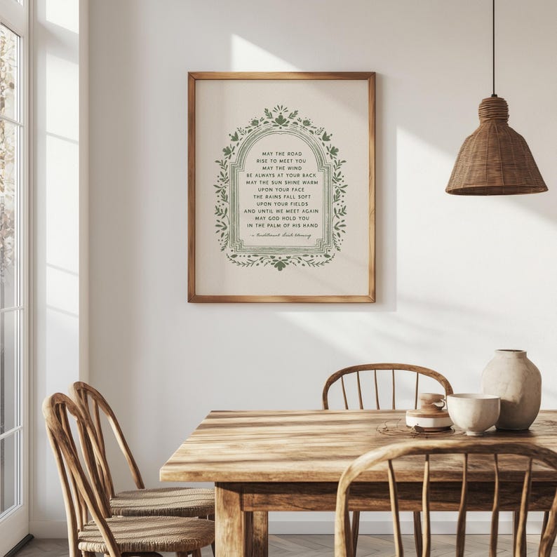 May include: A framed print with a wooden frame, featuring a green floral border and text. The text reads "May the Road Rise to Meet You." The print is displayed in a dining room setting with a wooden table and chairs.