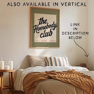 May include: A framed poster with the text "the Homebody club" in black script, above the words "LIVE LAUGH LOUNGE" in a red font. The poster hangs above a bed with pillows and a brown blanket. Candles sit on a wooden side table.
