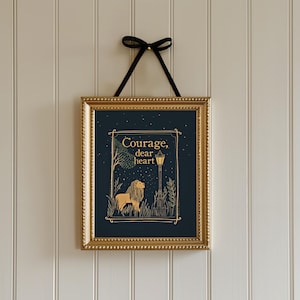 May include: A framed art print with a gold frame and a black ribbon for hanging. The artwork features a lion illustration with the words "Courage, dear heart" in gold lettering against a dark blue background. The print is displayed on a white paneled wall.