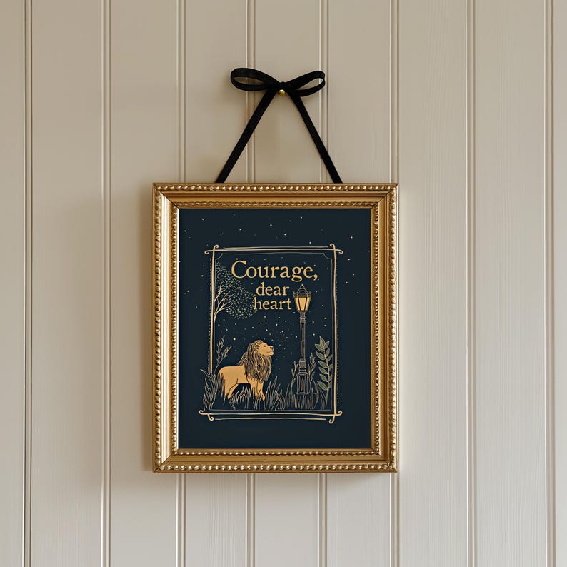 May include: A framed art print with a gold frame and a black ribbon for hanging. The artwork features a lion illustration with the words "Courage, dear heart" in gold lettering against a dark blue background. The print is displayed on a white paneled wall.