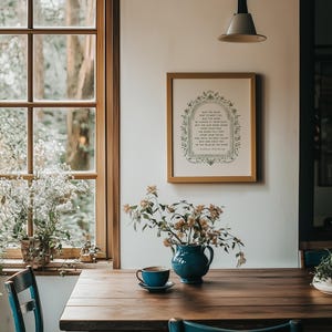 May include: A dining room scene featuring a wooden table with a blue ceramic pitcher holding flowers and a teacup. A framed print with text hangs on the wall. Blue chairs and a window with a view of trees complete the setting.