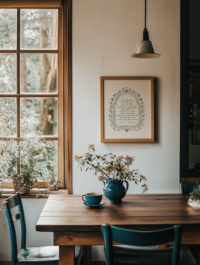 May include: A dining room scene featuring a wooden table with a blue ceramic pitcher holding flowers and a teacup. A framed print with text hangs on the wall. Blue chairs and a window with a view of trees complete the setting.