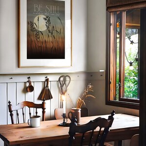 May include: A rustic wooden table with two chairs, a white mug, and a framed print with the text "Be Still and know that I am God". The print features a moon and tall grass in a muted color palette.