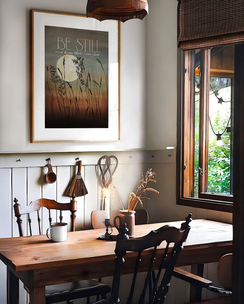 May include: A rustic wooden table with two chairs, a white mug, and a framed print with the text "Be Still and know that I am God". The print features a moon and tall grass in a muted color palette.