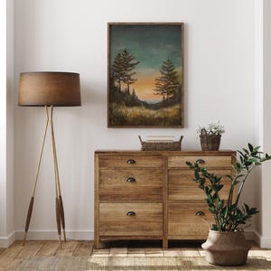 May include: A framed painting of a forest scene with two tall trees silhouetted against a sunset sky with stars. The painting is hanging on a white wall above a wooden dresser with drawers.