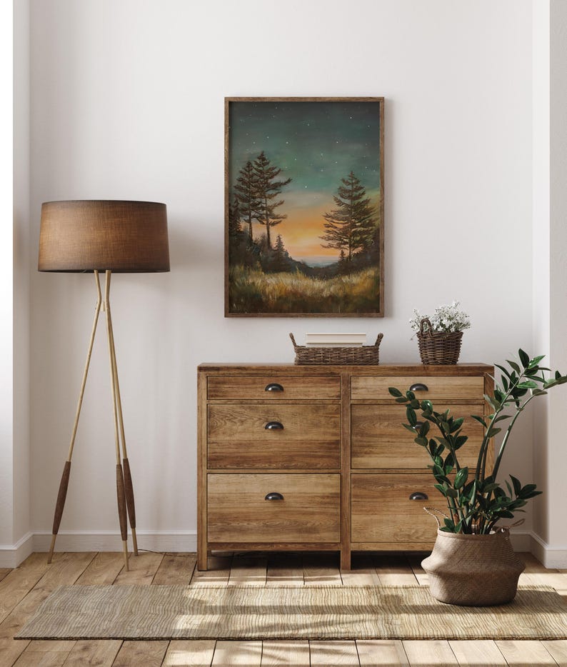 May include: A framed painting of a forest scene with two tall trees silhouetted against a sunset sky with stars. The painting is hanging on a white wall above a wooden dresser with drawers.