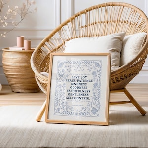 May include: A framed print with a blue floral border and white background. The text inside the frame reads "Love, Joy, Peace, Patience, Kindness, Goodness, Faithfulness, Gentleness, Self Control." The print is sitting on a white rug in front of a wicker chair.