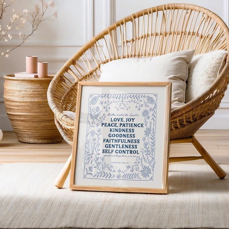 May include: A framed print with a blue floral border and white background. The text inside the frame reads "Love, Joy, Peace, Patience, Kindness, Goodness, Faithfulness, Gentleness, Self Control." The print is sitting on a white rug in front of a wicker chair.