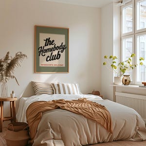 K&ouml;nnte beinhalten: Ein Schlafzimmer mit einem Bett mit beiger Bettw&auml;sche und einer braunen Decke. Ein gerahmtes Poster mit dem Text "the Homebody Club" h&auml;ngt an der Wand. Gestreifte Kissen, ein Holzschemel und eine Vase mit getrockneten Pflanzen erg&auml;nzen die Einrichtung.