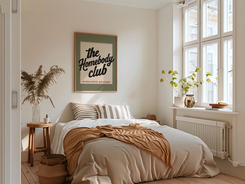 K&ouml;nnte beinhalten: Ein Schlafzimmer mit einem Bett mit beiger Bettw&auml;sche und einer braunen Decke. Ein gerahmtes Poster mit dem Text "the Homebody Club" h&auml;ngt an der Wand. Gestreifte Kissen, ein Holzschemel und eine Vase mit getrockneten Pflanzen erg&auml;nzen die Einrichtung.