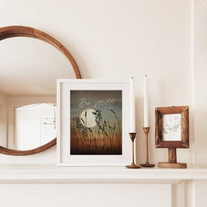 May include: A white framed print with a watercolor illustration of tall grass and a full moon. The text "Be still and know that I am God" is written in a cursive font.