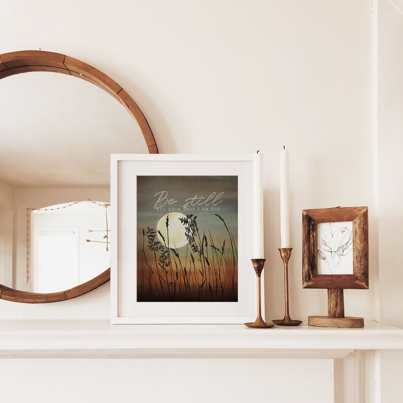 May include: A white framed print with a watercolor illustration of tall grass and a full moon. The text "Be still and know that I am God" is written in a cursive font.