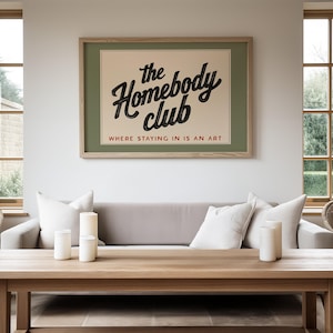 May include: A framed poster with the text "the Homebody club" in a black script font, with the tagline "WHERE STAYING IN IS AN ART". The poster is mounted above a gray sofa with white pillows, and a wooden coffee table.