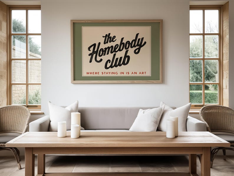 May include: A framed poster with the text "the Homebody club" in a black script font, with the tagline "WHERE STAYING IN IS AN ART". The poster is mounted above a gray sofa with white pillows, and a wooden coffee table.