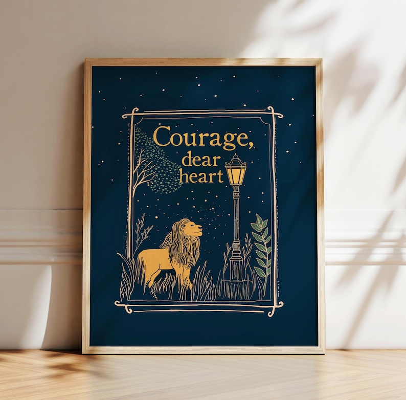 May include: A framed art print with a navy blue background. The artwork features a lion, a lamppost, and the words 'Courage, dear heart' in gold. The lion is depicted in a golden color, standing in a field of grass. The lamppost is black with a yellow light.