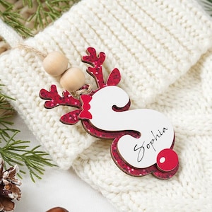 Peut inclure: Une chaussette de Noël en tricot de couleur crème avec une étiquette de nom de renne décorative. L'étiquette de renne est rouge et blanche avec le nom "Sophia" écrit dessus. L'étiquette est ornée de perles en bois et d'une boule rouge.