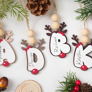 May include: Wooden Christmas ornaments shaped like reindeer with names on them. Each ornament has a letter or a letter-shaped design, a red bow, and a red ball. The ornaments are hanging from a string with wooden beads.
