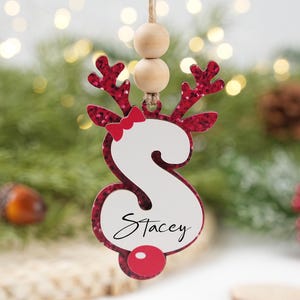 Puede incluir: Adorno navideño con forma de reno con acabado de purpurina roja. El adorno presenta la letra "S" con el nombre "Stacey" en escritura cursiva. Un lazo y una nariz rojos completan el diseño, con cuentas de madera en la parte superior para colgar.