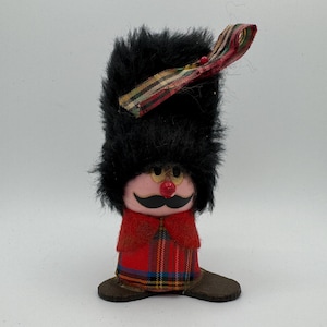 Pode incluir: Estatueta decorativa de um soldado com um chapéu preto peludo, um rosto rosa com um bigode preto e uma roupa vermelha e xadrez. A estatueta tem um nariz vermelho e sapatos castanhos. Uma fita colorida está presa ao chapéu.
