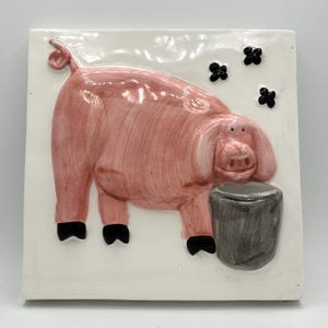 May include: A pink ceramic tile with a 3D image of a pig with a curled tail, standing on all four legs and looking at a gray bucket. The background is white with black flower shapes.