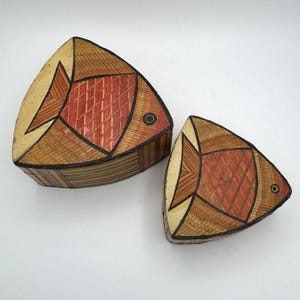 Op de afbeelding: Twee houten, visvormige dozen met geometrische patronen in tinten van bruin, rood en zwart. De grotere doos is op de voorgrond en de kleinere doos is erachter.
