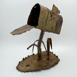Op de afbeelding: Een roestig metalen sculptuur van een brievenbus met een vlag erop. De brievenbus is bevestigd aan een metalen basis met drie metalen stengels die op gras lijken.