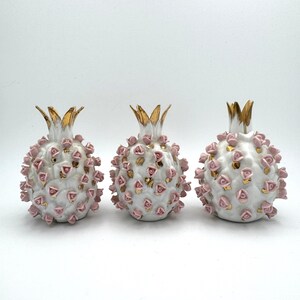 May include: Three white ceramic pomegranate sculptures with gold crowns and accents. Each pomegranate is decorated with small pink rose-shaped embellishments. The sculptures are arranged on a white surface.