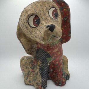 May include: A vintage paper mache dog figurine with large eyes and a patchwork design. The dog is sitting with its head tilted slightly to the side. The patchwork design features floral patterns in shades of red, blue, and green.