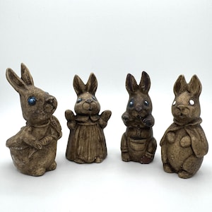 May include: Four brown ceramic bunny figurines with blue eyes. The bunnies are wearing different outfits and have different facial expressions.