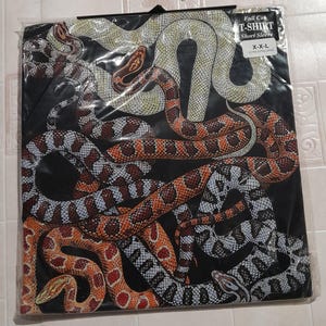 Puede incluir: Camiseta negra con un estampado gráfico de varias serpientes entrelazadas. Las serpientes son de color naranja, marrón, blanco y negro. La camiseta es de corte completo, manga corta y talla extra grande.
