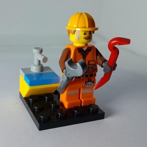 May include: A Lego minifigure of a construction worker wearing an orange vest and yellow hard hat. The figure holds a gray wrench and a red crowbar. The figure stands on a black base with a small structure.