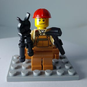 Può includere: Una minifigure LEGO che indossa un elmetto rosso e una salopette color marrone chiaro con un design a tasca stampato. Il personaggio ha una testa gialla con un sorriso, tiene un attrezzo nero in ogni mano e poggia su una base LEGO grigia.