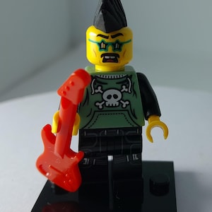 May include: A Lego minifigure with a black mohawk hairstyle, yellow skin, and star-shaped glasses. The figure wears a light green hoodie featuring a white skull and crossbones graphic, black pants, and holds a bright red electric guitar accessory.