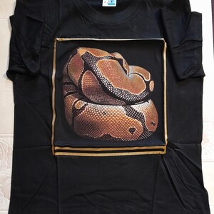Puede incluir: Camiseta negra con una imagen enmarcada en oro de una serpiente marrón y negra.