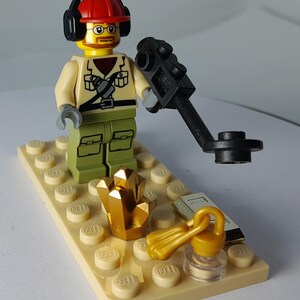 May include: A Lego figurine of an archaeologist wearing a red hard hat, glasses, and headphones, holding a black metal detector. The scene includes a gold crystal, a small book, and a gold lantern on a tan Lego base.