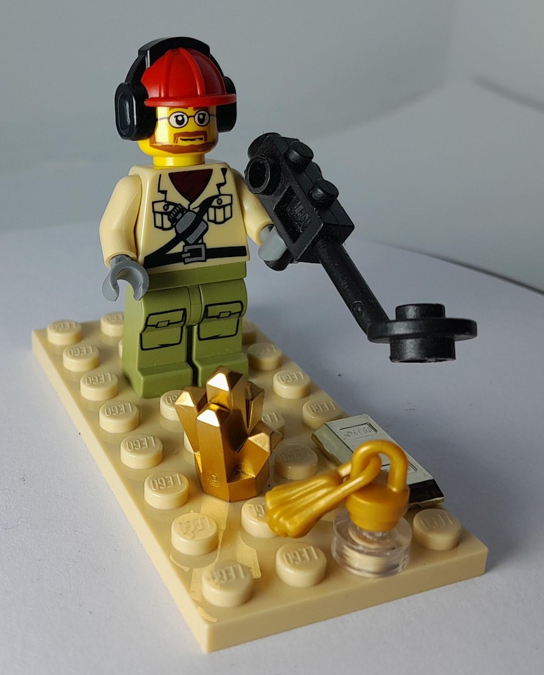 May include: A Lego figurine of an archaeologist wearing a red hard hat, glasses, and headphones, holding a black metal detector. The scene includes a gold crystal, a small book, and a gold lantern on a tan Lego base.