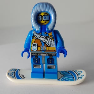 Könnte beinhalten: Eine Lego-Minifigur in einem blauen Winteroutfit mit einer fellgefütterten Kapuze und Brille, die auf einem weißen und blauen Snowboard steht. Das Snowboard hat ein Design und den Text "48mm". Die Minifigur hat ein detailliertes Torso-Design.