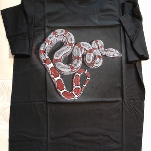 Può includere: Maglietta nera con stampa grafica di un serpente rosso e bianco.