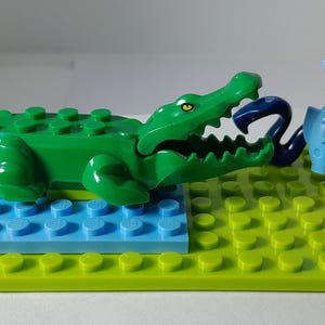 Op de afbeelding: Een groen plastic krokodillenspeeltje met een open bek, een donkerblauwe slang en een lichtblauwe vis vastgehouden door een blond minifiguur in een beige shirt en broek. De figuren staan op een limoengroene basis met een blauwe plaat.