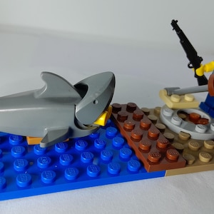 Puede incluir: Una construcción de LEGO con una figura de tiburón gris sobre una placa azul que representa agua, junto a una plataforma marrón. Una minifigura con cabello negro, camisa azul y pantalones azules sostiene un rifle negro y una vara gris larga.