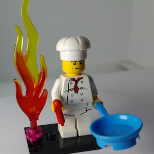 May include: A Lego chef minifigure in a white uniform and chef's hat, holding a red sausage and a blue frying pan. A flame accessory with red and yellow hues stands beside the chef on a black base. The chef has a red tie.