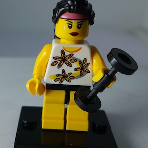 以下が含まれることがあります： 黄色い肌、黒い髪、ピンクのヘッドバンドを身につけたLEGOミニフィギュア。オレンジと茶色の花柄が描かれた白いタンクトップと黄色い脚部を着用している。黒いプラスチック製のダンベルを持ち、黒い正方形の台座の上に立っている。