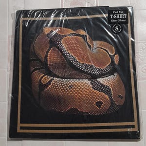 Puede incluir: Una camiseta negra con un gráfico de una serpiente marrón y negra. La camiseta tiene un borde dorado y el texto "Full Cut T-SHIRT Short Sleeve" en blanco. La talla "S" también está impresa en la camiseta.