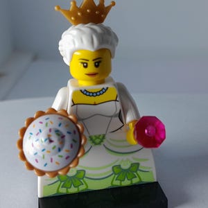 May include: A Lego minifigure wearing a white gown with green bow accents and a gold crown. The figure has white hair, a yellow face, and a blue necklace. It holds a round cupcake-shaped accessory with colourful sprinkles and a pink gem piece.