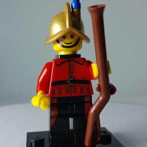 May include: A Lego minifigure of a soldier in a red jacket, black trousers, and a gold helmet with a blue plume. The figure holds a brown rifle and stands on a black base. The figure has a smiling face.