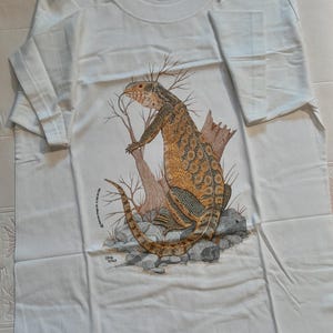 Puede incluir: Camiseta blanca con una ilustración de un lagarto marrón y amarillo. El lagarto está de pie sobre rocas y ramas. El texto "Leon Hill" está impreso en la parte inferior de la camiseta.